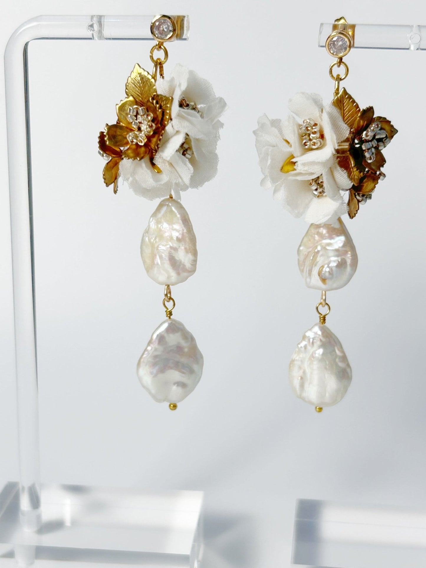 Pair of earrings with floral and pearl designs on a clear stand.