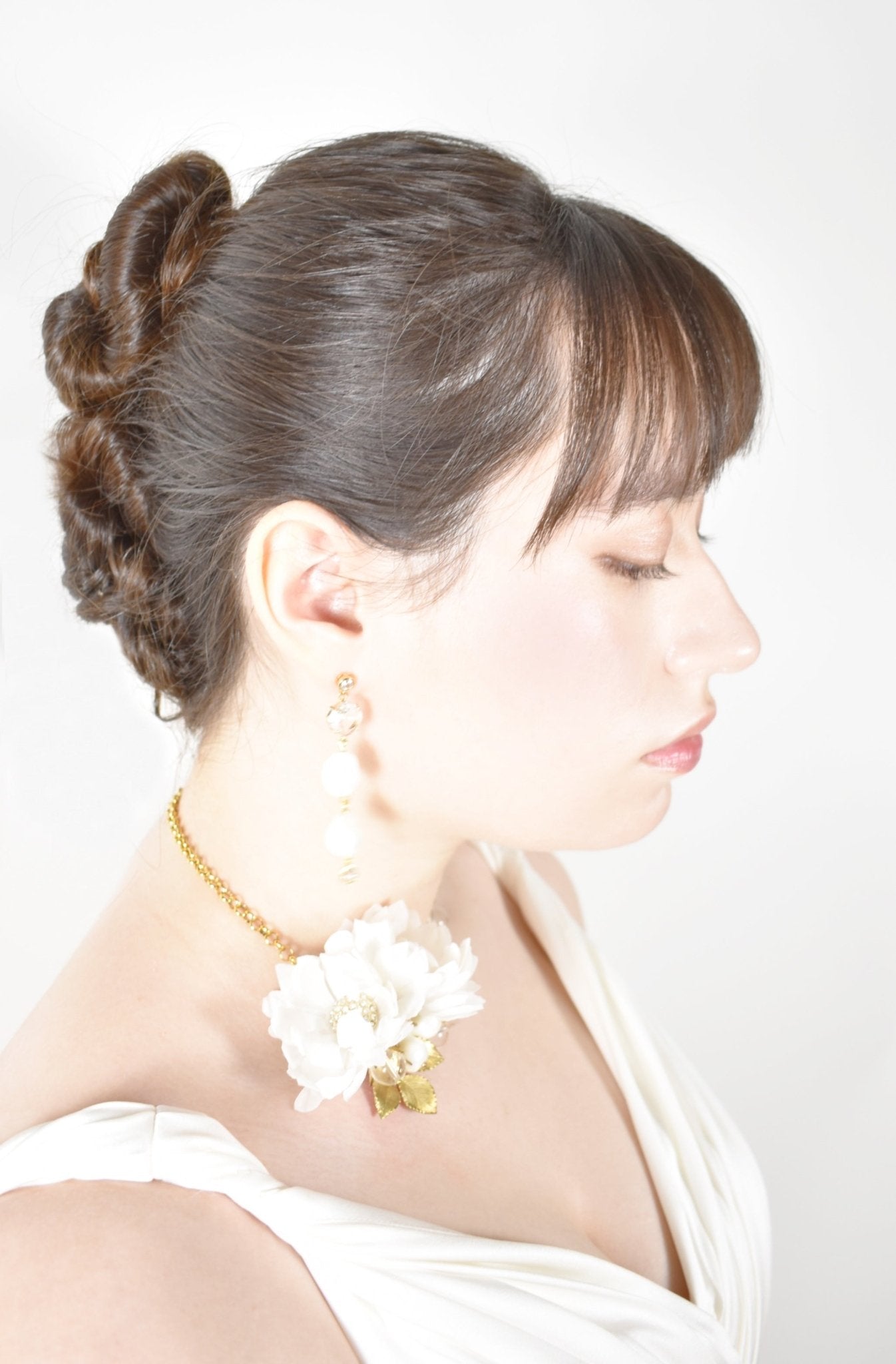 Woman with styled hair and white flowers, wearing a gold earring and necklace, on a plain background