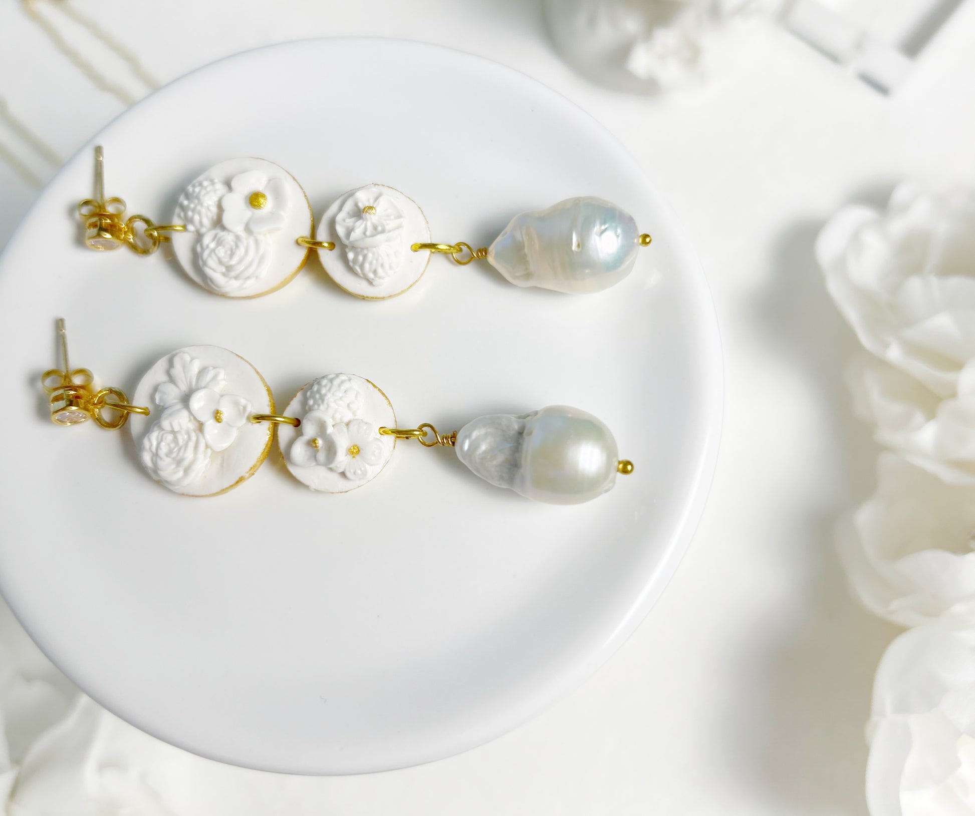 Pair of earrings with white floral designs and pearl-like beads on a white plate.
