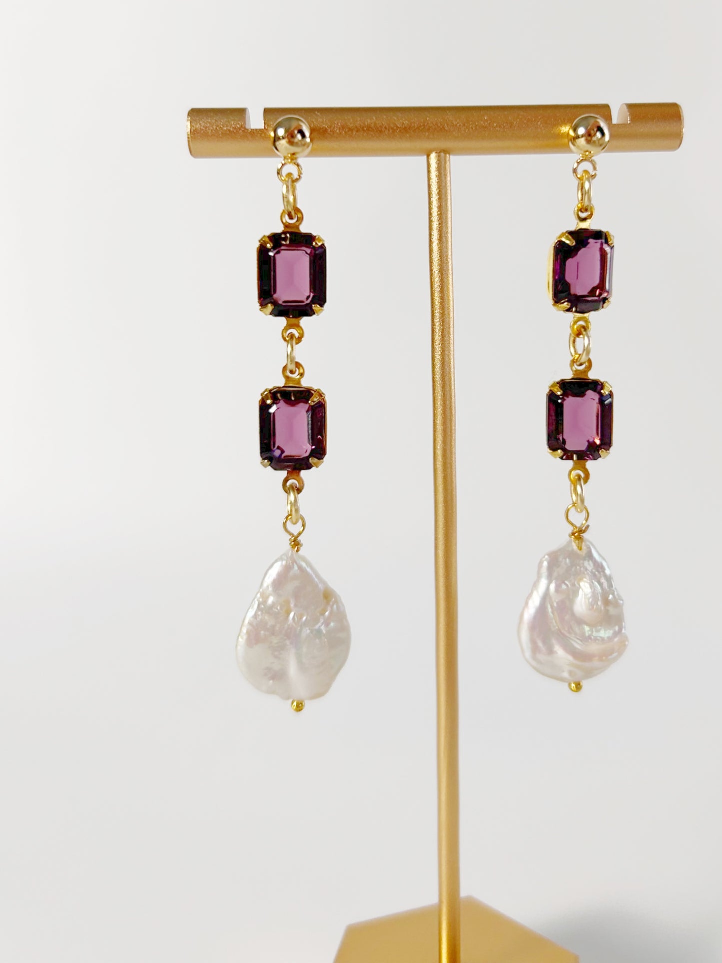 Gold earrings with purple gemstones and pearl-like drops on a gold stand against a white background